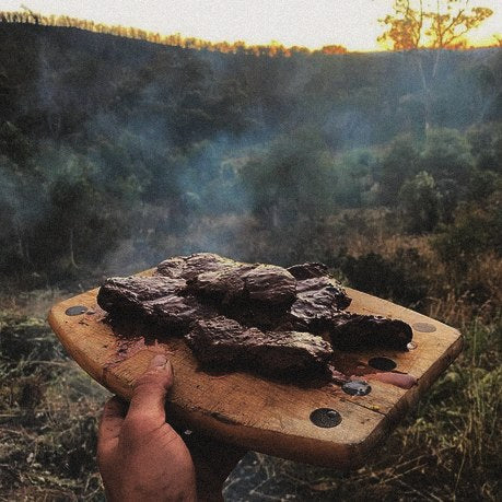 A hand holding a wooden board of fire-cooked meat over a wooded valley at sunset